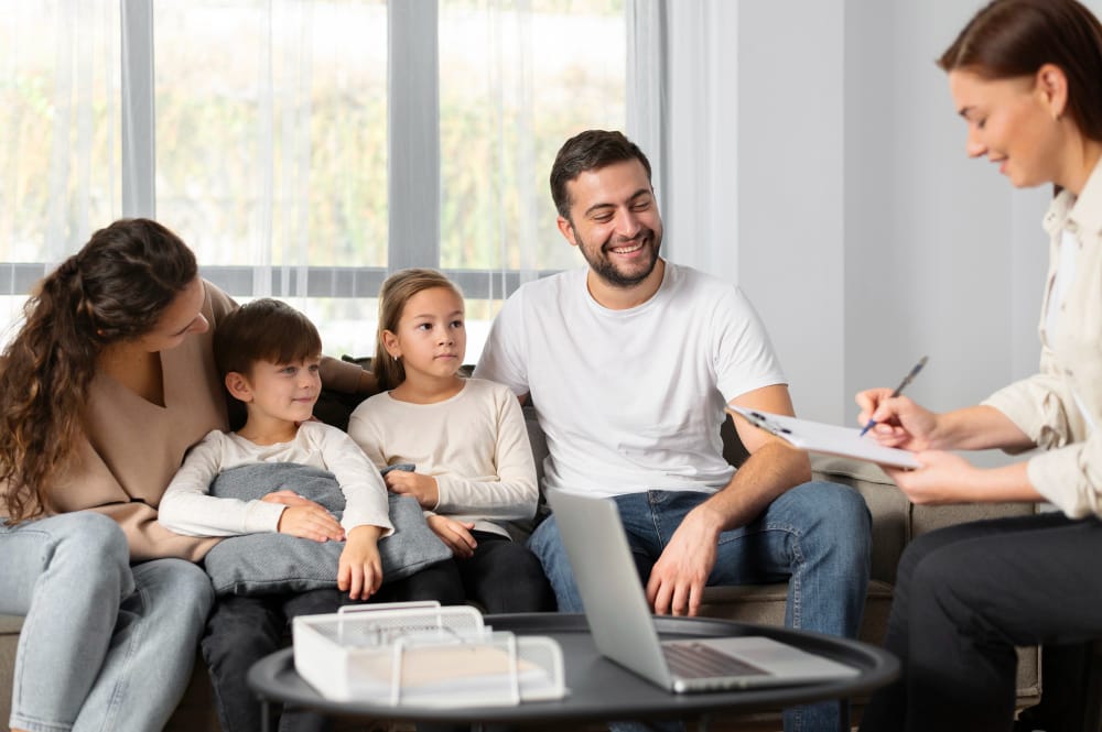 A smiling family sits together on a sofa while a female consultant takes notes on a clipboard, she’s writing down that she will bill this as 90846 CPT code services. The family includes a man, a woman, and two children, all appearing engaged and relaxed. A laptop is placed on the coffee table in front of them.