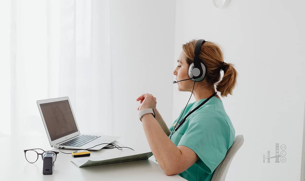 A woman in a teal-colored scrub top and headphones, a medical professional, is seated at a desk, using a laptop. The image is about telehealth & remote consultation scenarios.