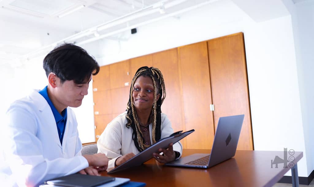 Two medical professionals are seated at a table, interacting and working. They are reviewing CPT code 98967 billing documents or discussing. A laptop is visible on the table.