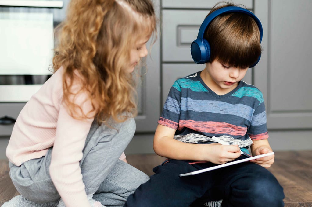 One child is concentrated on his tablet and has headphones on while the other tries to talk to him.