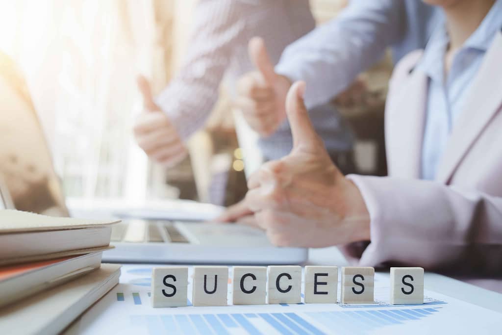  Professionals give a thumbs up in a teamwork gesture over a desk with documents and devices, symbolizing collaboration and implementation of best practices in a business setting.