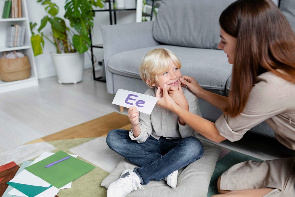 Young woman conducting a speech therapy session with a young blonde boy, using educational tools in a bright, supportive environment. The scene represents early intervention for a child diagnosed with F84.0 (Autistic Disorder).