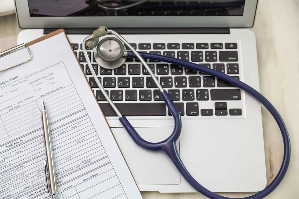 Top view of a laptop on a desk with a stethoscope placed on the keyboard and health records.