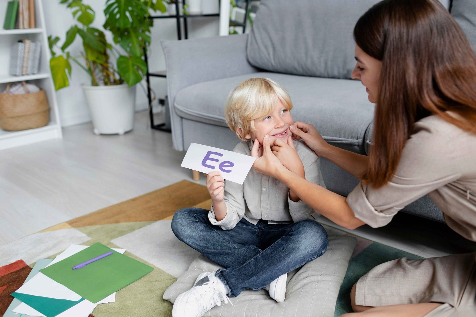 Young woman conducting a speech therapy session with a young blonde boy, using educational tools in a bright, supportive environment. The scene represents early intervention for a child diagnosed with F84.0 (Autistic Disorder).